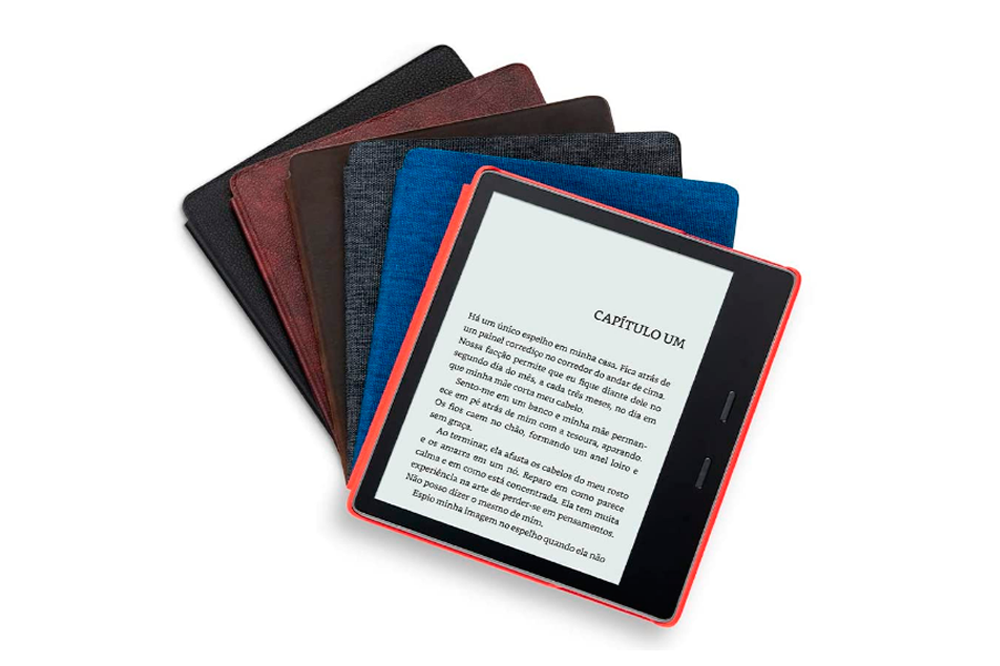 A imagem mostra vários kindles empilhados, com diferentes cores de capinhas: preto, vermelho, marrom, cinza, azul e, no topo de todos, há um kindle aberto, mostrando sua tela com o capítulo de um livro no fundo branco. É possível ver que sua capinha é laranja. O fundo da imagem é branco.