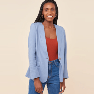 A imagem mostra uma mulher negra, com cabelos compridos em tranças, usando um blazer de linho azul claro, com uma regata na cor terra embaixo do blazer. Ela usa calça jeans e está num fundo rosa claro.