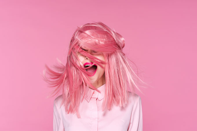 Mulher com peruca rosa, à frente de parede rosa, e camisa branca, com lábios vermelhos