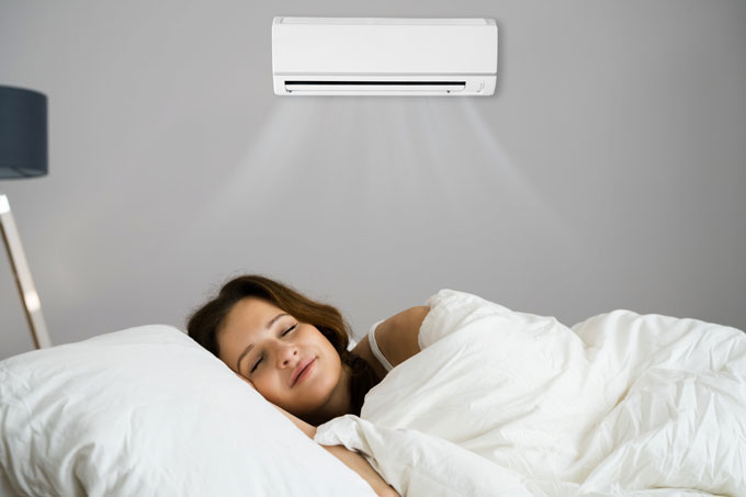 Mulher dorme em sua cama, no primeiro plano, com um aparelho de ar-condicionado na parede atrás