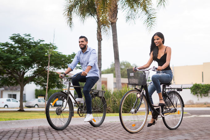 Casal andando de bicicletas em uma calçada ampla, com coqueiros ao fundo