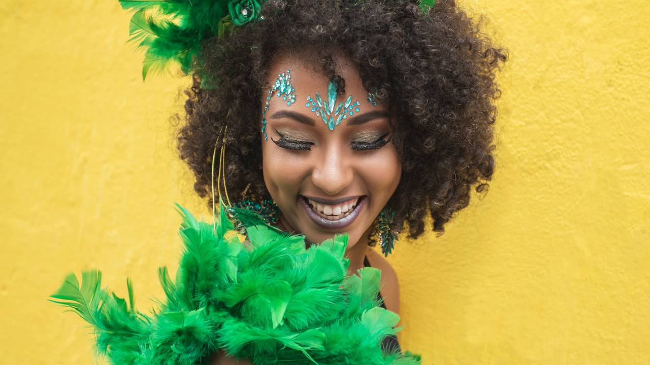 mulher negra com cabelo black power, fantasiada com penas verdes, de maquiagem azul e em fundo amarelo