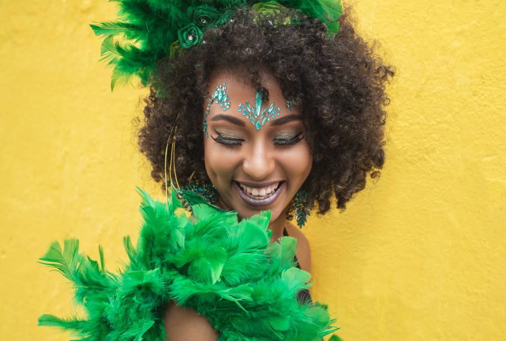 mulher negra com cabelo black power, fantasiada com penas verdes, de maquiagem azul e em fundo amarelo